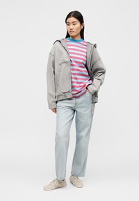 Γυναίκα με γκρι υπερμεγέθη μπουφάν, μπλε και ροζ ριγέ πουκάμισο, ανοιχτό μπλε τζιν και μπεζ αθλητικά παπούτσια, στέκεται με τα χέρια στις τσέπες.