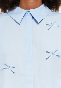 Camisa de colarinho azul-claro com pequenos laços de tecido decorados com contas. Textura suave e fecho de botão frontal.