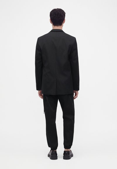 Homme debout, de dos, portant un blazer noir, un pantalon noir et des chaussures noires sur un fond blanc uni.