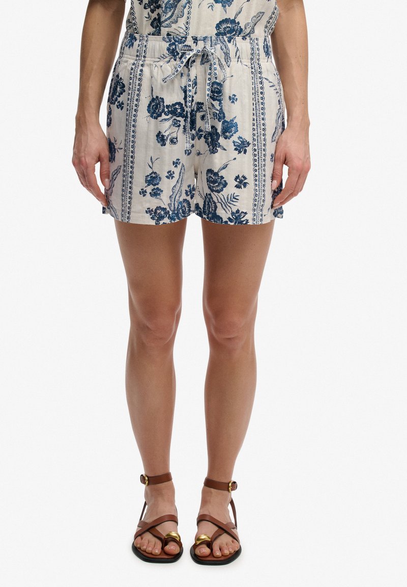 Mujer vistiendo pantalones cortos beige claro con estampado floral azul y cintura anudada, combinados con sandalias planas marrones con tiras en el tobillo.