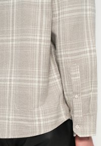 Manga larga a cuadros beige y blancos con puño de dos botones blancos, combinado con pantalones negros, mostrando detalles de los lados y las mangas.
