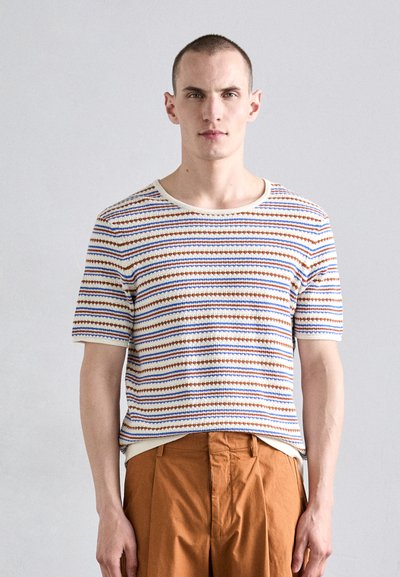 Gestreepte gebreide shirt met crème, blauwe en roestkleurige lijnen; ronde hals; korte mouwen. Gedragen met bruine, geplooide broeken.