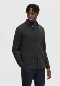 Camisa negra de botones en un tejido texturizado, con cuello clásico y cierre de botones. Lleva puesta sobre un suéter de punto azul marino con patrón, combinado con pantalones de cuadros.