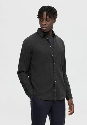 Camisa negra de botones en un tejido texturizado, con cuello clásico y cierre de botones. Lleva puesta sobre un suéter de punto azul marino con patrón, combinado con pantalones de cuadros.
