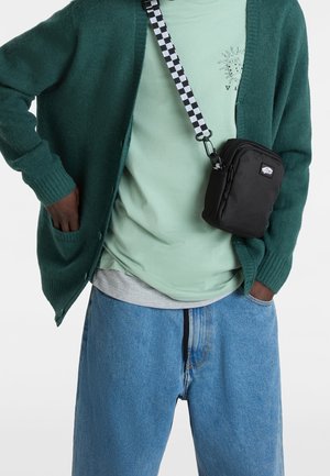 Persona que lleva un cárdigan color verde azulado y una camiseta gráfica verde menta, con jeans azules holgados, llevando un bolso cruzado negro con correa de cuadros.