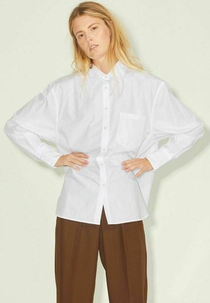 Camisa blanca de manga larga con botones y un bolsillo. Hecha de tela suave, con un corte relajado y puños anchos. Combinada con pantalones marrones.