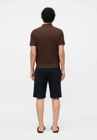 Άνδρας με σγουρά μαλλιά στέκεται πλάτη, φοράει καφέ κοντομάνικο πουκάμισο, μαύρα σορτς μέχρι το γόνατο και μαύρα σανδάλια slide σε λευκό φόντο.