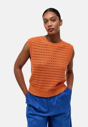Pull en crochet orange avec un motif texturé et des bordures côtelées. Design sans manches associé à un pantalon bleu, créant un look contrasté.