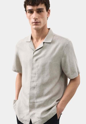 Junger Mann trägt ein hellbeiges, kurzärmeliges Hemd mit Kragen und Knöpfen, Hände in den Taschen, neutraler Gesichtsausdruck, schlichter heller Hintergrund.