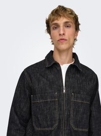 Veste en denim indigo foncé avec fermeture éclair frontale, deux poches poitrine, et surpiqûres contrastantes orange. Tissu texturé, col structuré.