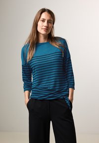 Turbaanin pitkät hiharesorvinen tee jossa on tummansinisiä vaaka-raitoja ja pyöreä pääntie. Vyötäröllä on solmittu yksityiskohta ja rento istuvuus. Mustat housut.