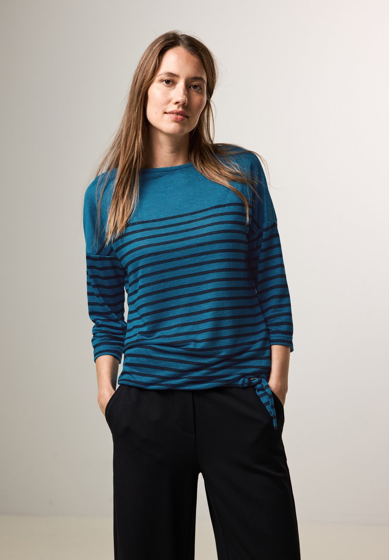 Turbaanin pitkät hiharesorvinen tee jossa on tummansinisiä vaaka-raitoja ja pyöreä pääntie. Vyötäröllä on solmittu yksityiskohta ja rento istuvuus. Mustat housut.