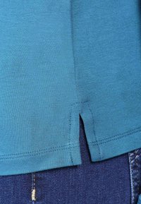 Türkises kurzärmliges Shirt mit geradem Saum und seitlichen Schlitzen, aus weichem Stoff, kombiniert mit dunkelblauen Jeans mit sichtbaren Nähten.