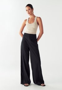 Bež majica z ribanim vzorcem in visoko pasne črne široke hlače. Hlače imajo pregledne gube; model nosi črne sandale z zlatimi detajli.