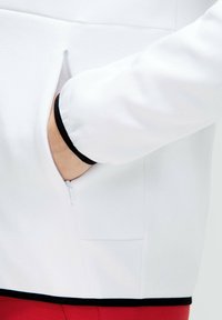 Indumento in tessuto bianco con tasca laterale con zip e rifiniture nere all'orlo e alle estremità delle maniche, che presenta una texture liscia.