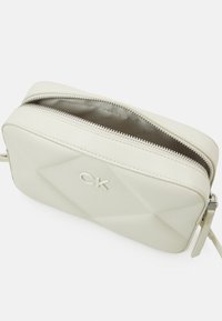 Bolso cruzado de cuero sintético color crema con textura de diamante, cierre superior de cremallera y logotipo de CK en relieve en la parte frontal.