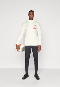 Muški trenirka u krem boji s crvenim Liverpool logotipom, uparen s crnim hlačama i odgovarajućim crnim nogometnim cipelama, drži uzorčanu nogometnu loptu.