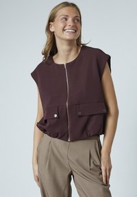 Bordeaux zip-up vest med korte ærmer, der har to forlommer og knapdetaljer. Matchet med beige bukser med høj talje.