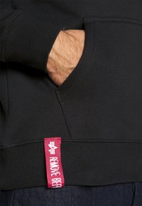 Felpa nera con tasca frontale, polsini a costine e un'etichetta rossa con logo sull'orlo con testo bianco. Il tessuto appare morbido.