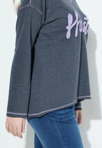 Maglia grigia a maniche lunghe con testo ricamato in viola, orlo curvo e cuciture a contrasto, abbinata a jeans in denim blu.