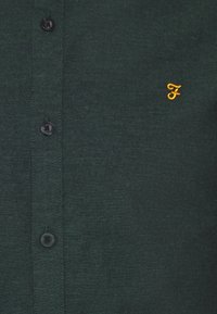 Chemise boutonnée vert foncé avec trois boutons noirs visibles et petite lettre "F" jaune brodée sur la poitrine.