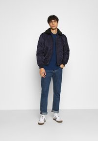 Marineblaue Bomberjacke mit schwarzem Fellkragen, getragen über einem blauen Poloshirt, kombiniert mit hochgekrempelten dunkelblauen Jeans und weißen Sneakers.