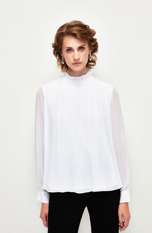 Blouse blanche plissée avec un col haut à volants et des manches longues transparentes, portée sur un pantalon noir. Texture lisse avec un design fluide.