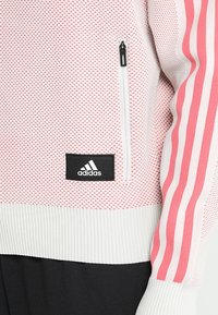 Sudadera ligera de malla con rayas alternas en rosa y blanco, bolsillo con cremallera y parche del logo de adidas en negro en el dobladillo. Tejido texturizado.