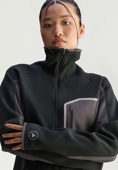 Giacca nera con zip e colletto alto, realizzata in tessuto strutturato, con accenti grigi e una tasca con zip sul petto.