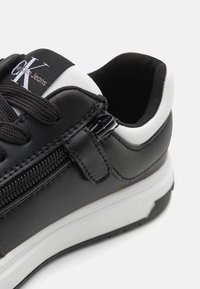 Sneaker in pelle nera con accenti bianchi, punta tonda, superficie testurizzata e chiusura laterale con zip; presenta un colletto imbottito e lacci neri.