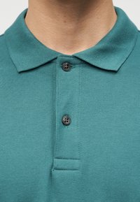 Gros plan sur le col d'un polo bleu sarcelle avec deux boutons noirs attachés sur le cou et le haut de la poitrine d'une personne.