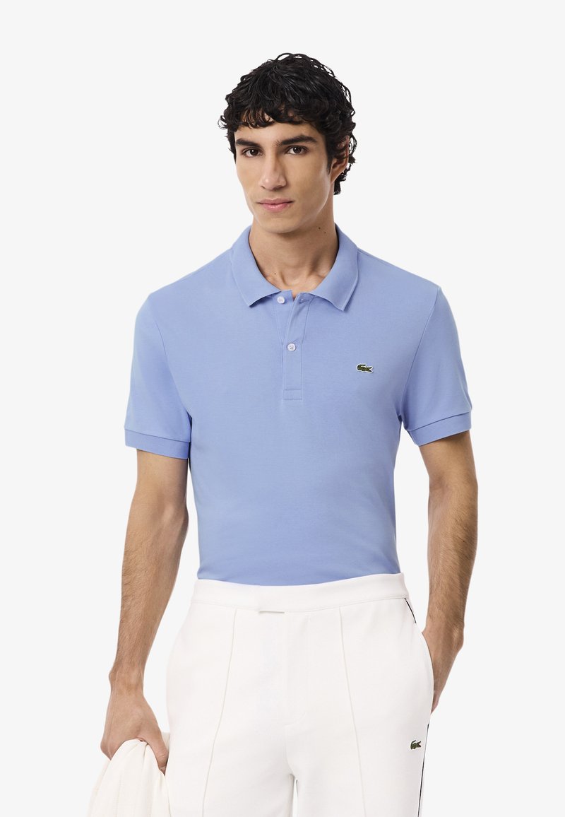 Polo shirt azzurro chiaro realizzato in tessuto morbido, con colletto, maniche corte e un dettaglio del logo verde sul petto. Indossato con pantaloni bianchi.