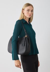 Donna con una blusa verde acqua e pantaloni neri porta a spalla una borsa nera testurizzata con dettagli a frangia.