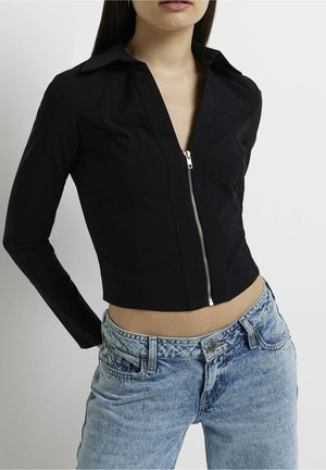 Kvinde iført en tætsiddende sort cropped jakke med lange ærmer, frontlynlås og lyseblå højttaljede jeans.