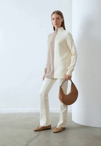 Beige and cream color-blocked oversized sweater with high neck, paired with white fitted trousers and a brown crescent bag. Flat shoes.