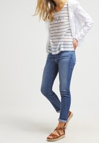 T-shirt rigato bianco e blu navy sovrapposto a una giacca in pizzo bianca, abbinato a jeans skinny blu chiaro e sandali con cinturini marroni.