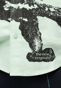 Lys grøn skjorte med knapper, med sort pixelagtigt abstrakt design og hvid tekst "the new originals" nær bunden af kanten.