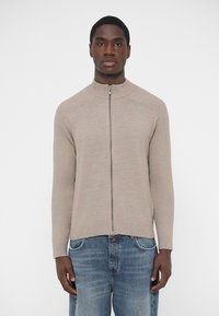 Svetlohnedý sveter na zips s rebrovanou textúrou, vysokým golierom a dlhými rukávmi, v kombinácii s modrými džínsami. Má hladký povrch a priliehavý strih.