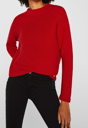 Frau trägt einen roten, langärmeligen, gerippten Pullover und schwarze Hosen, steht mit einer Hand auf der Hüfte.