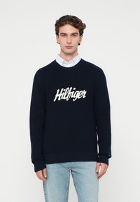 Granatowy sweter z dzianiny z okrągłym dekoltem i ściągaczami, z wyraźnym białym logo "Hilfiger" na klatce piersiowej.