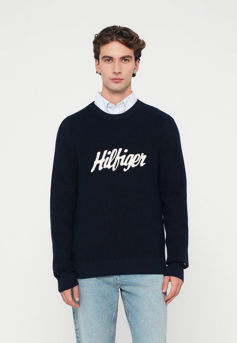Granatowy sweter z dzianiny z okrągłym dekoltem i ściągaczami, z wyraźnym białym logo "Hilfiger" na klatce piersiowej.