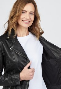 Svart lädermotorcykeljacka med silverdetaljer, dragkedjestängning och sidofickor, bärs över en vit skjorta. Slät textur, skräddarsydd passform.