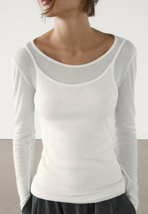 T-shirt blanc à manches longues avec un design superposé ; il est fabriqué dans un tissu léger et doux, et possède un col rond avec une bordure contrastante.