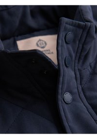 Colletto di giacca trapuntata blu navy con due bottoni rotondi blu. Texture morbida, cuciture visibili e un'etichetta interna.