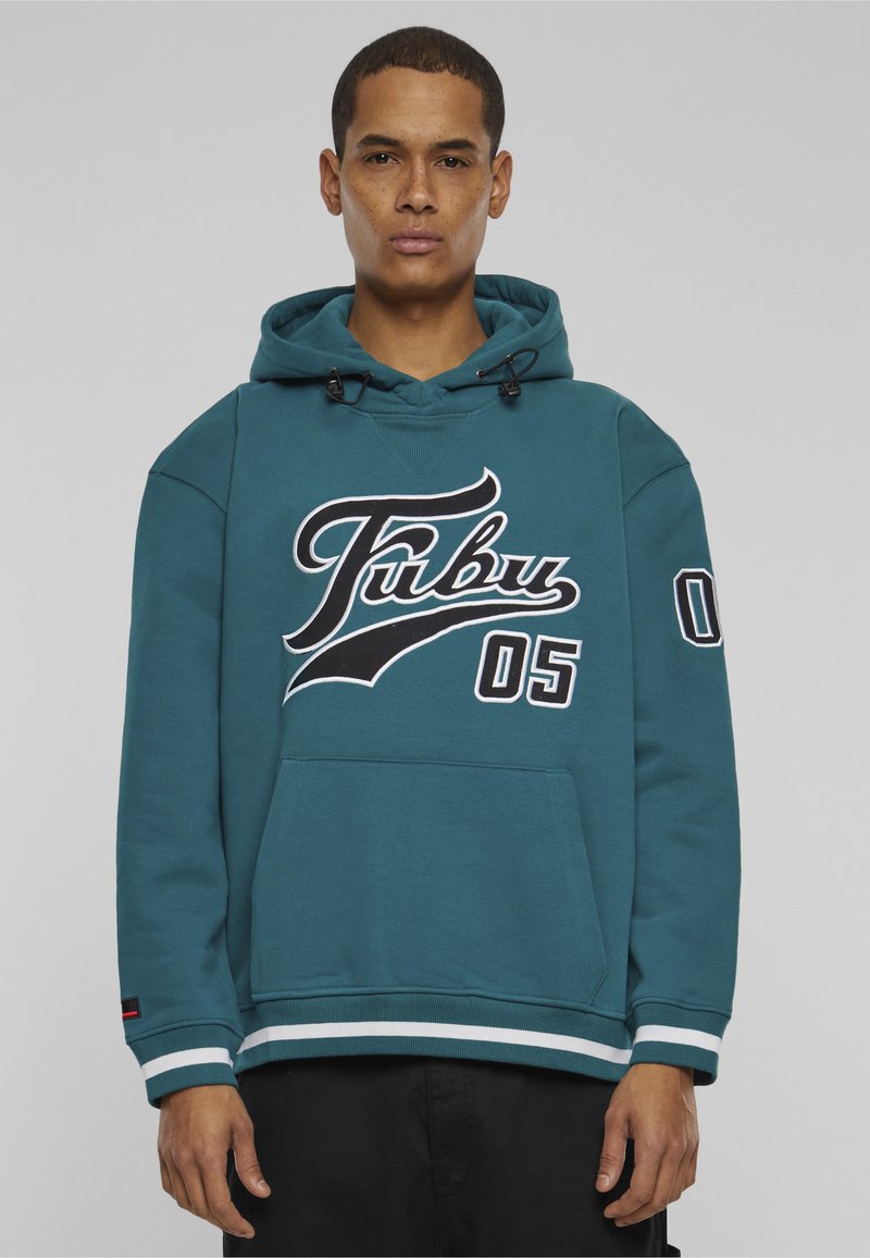 Junger Mann trägt einen türkisfarbenen Fubu-Hoodie mit weißen und schwarzen Buchstaben und der Zahl 05, vor einem schlichten grauen Hintergrund stehend.
