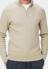 Beige ribstrikket sweater med høj hals og kvart-lynlås. Har lange ærmer og en tætsiddende silhuet. Blød tekstur.