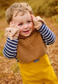 Rjav flis brezrokavnik z zadrgo preko mornarsko modre in bele prugaste srajce z dolgimi rokavi, rumene dežne hlače in svetlo rjave brezprstne rokavice z naglasi v obliki medveda.