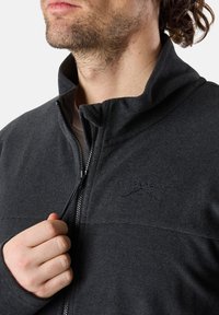 Svart fleecejacka med hög krage, framdragkedja och tonalt logotyp på bröstet. Materialet har en texturerad finish.