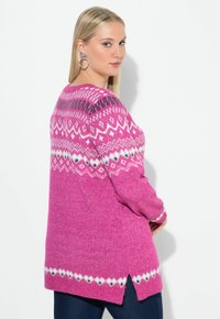 Kobieta w różowym swetrze w wzory z białymi i czarnymi motywami, stojąca bokiem z rękami w kieszeniach na białym, jednolitym tle.