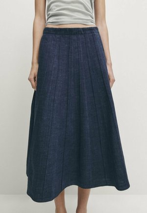 Woman wearing a high-waisted, dark blue, pleated midi skirt and a light gray sleeveless top, standing against a plain background.